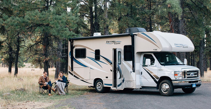 Two people sitting in folding chairs in front of an RV in the woods