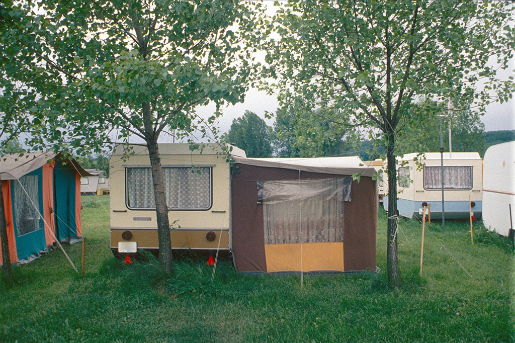Several vintage travel trailers and canvas tents set up on a grassy campground, flanked by trees. The trailers have retro designs and patterned curtains, suggesting a nostalgic camping scene.